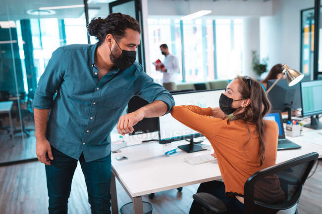 Two diverse businesspeople wearing face mask greeting with elbows and using computer. work at a modern office during covid 19 coronavirus pandemic. — Stock Photo