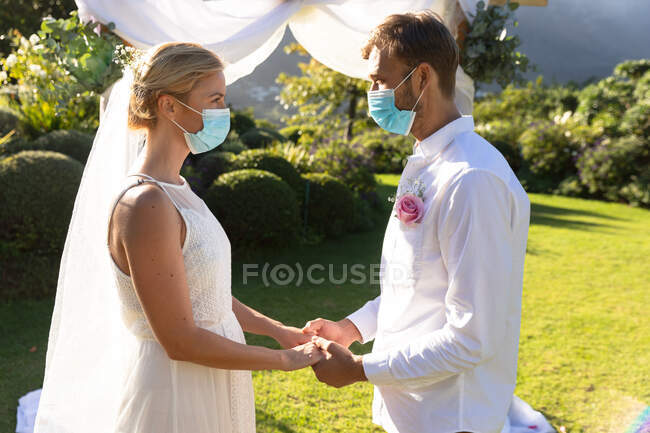 Heureux mariés caucasiens se marier portant des masques et se tenant la main. mariage d'été, mariage, amour et célébration pendant covid 19 concept de pandémie. — Photo de stock