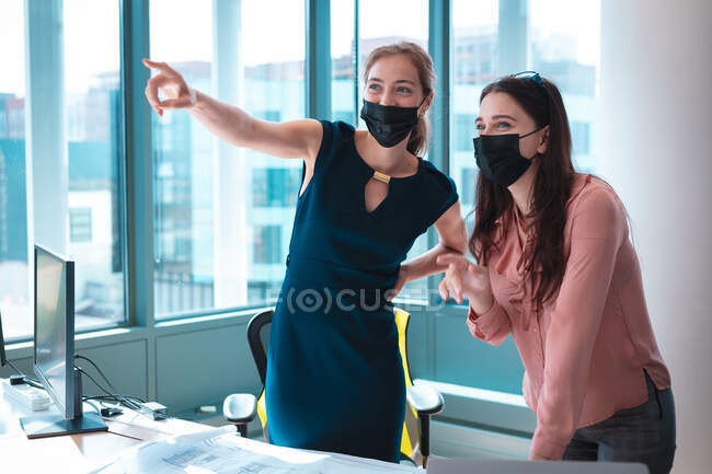 Dos empresarias caucásicas usando mascarilla y discutiendo. trabajar en una oficina moderna durante la pandemia de coronavirus covid 19. - foto de stock