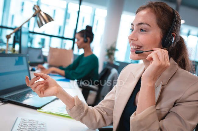 Kaukasische Geschäftsfrau mit Headset, die mit Kollegen im Hintergrund am Tisch sitzt. Arbeit in einem modernen Büro. — Stockfoto