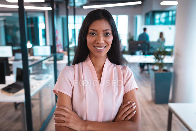 Retrato de mulher de negócios de raça mista sorrindo com braços cruzados com colegas de fundo. trabalho em um escritório moderno. — Fotografia de Stock