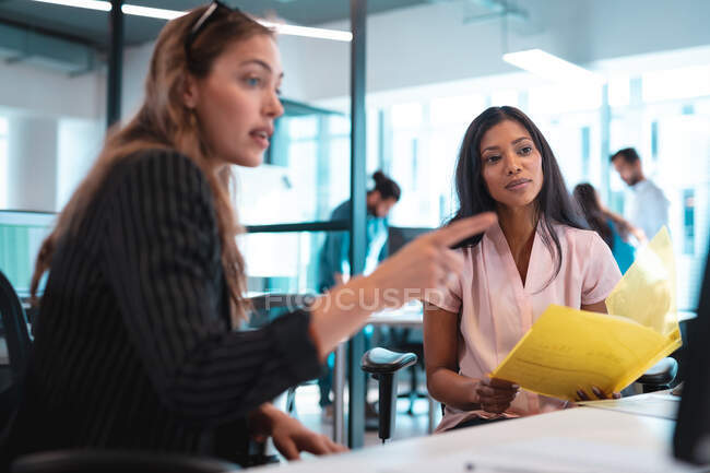 Due donne d'affari diverse che siedono a tavola e usano il computer con colleghi in background. lavorare in un ufficio moderno. — Foto stock