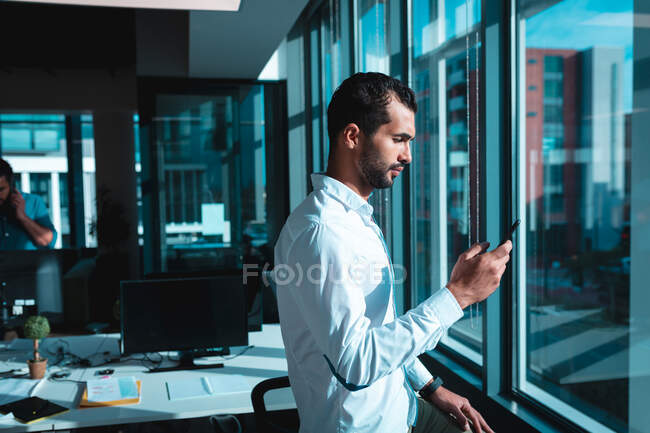 Gemischter Geschäftsmann am Fenster mit Smartphone und Kollegen im Hintergrund. Arbeit in einem modernen Büro. — Stockfoto