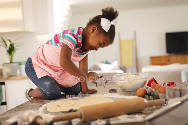 Feliz hija afroamericana arrodillada en el mostrador de la cocina cortando formas en masa. pasar tiempo libre en casa. - foto de stock