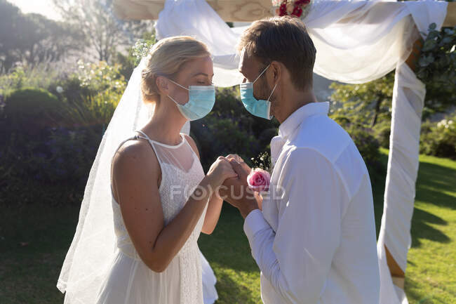 Felici sposi caucasici che si sposano indossando maschere facciali e tenendosi per mano. matrimonio estivo, matrimonio, amore e celebrazione durante covid 19 concetto pandemico. — Foto stock
