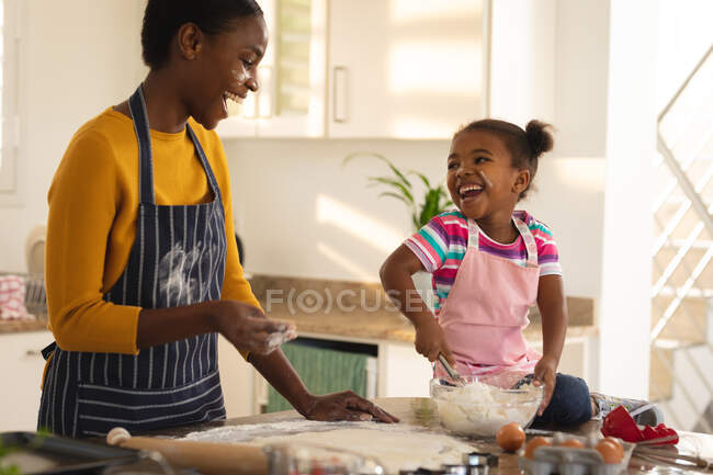 Смеющиеся африканская американская мать и дочь пекут вместе на кухне тесто. Семья проводит время вместе дома. — стоковое фото