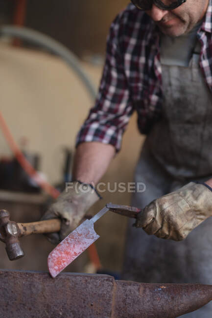 Primer plano de fabricante de cuchillo masculino caucásico usando delantal y gafas, utilizando martillo en el taller. artesano independiente de la pequeña empresa en el trabajo. - foto de stock