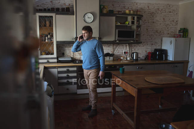 Фокусований білий чоловік стоїть на кухні, дивлячись у вікно, використовуючи смартфон. вільний час вдома . — стокове фото