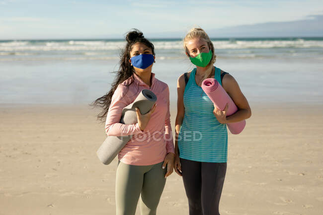 Due diverse amiche donne che indossano maschere che praticano yoga, in piedi, tenendo tappeti in spiaggia. sano stile di vita attivo, fitness all'aperto e benessere durante la pandemia della covd 19. — Foto stock