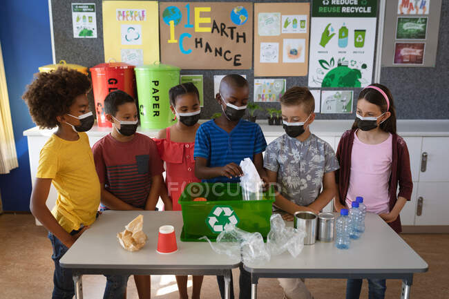 Groupe d'élèves divers portant des masques faciaux mettant des articles en plastique recyclable dans un plateau à l'école. éducation retour à l'école sécurité sanitaire pendant la pandémie de coronavirus covid19. — Photo de stock