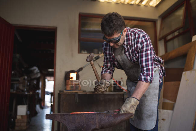 Caucasian male knife maker wearing apron and glasses, using hammer in workshop. independent small business craftsman at work. — Stock Photo