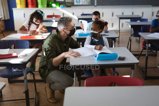 Maestra masculina caucásica usando mascarilla enseñando a una chica afroamericana en la clase de la escuela. higiene y distanciamiento social en la escuela durante la pandemia de covid 19 - foto de stock