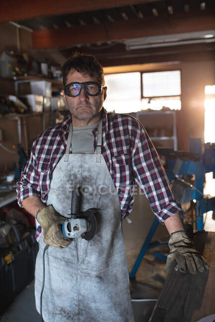 Caucasian male knife maker wearing apron and glasses, holding angle grinder in workshop. independent small business craftsman at work. — Stock Photo