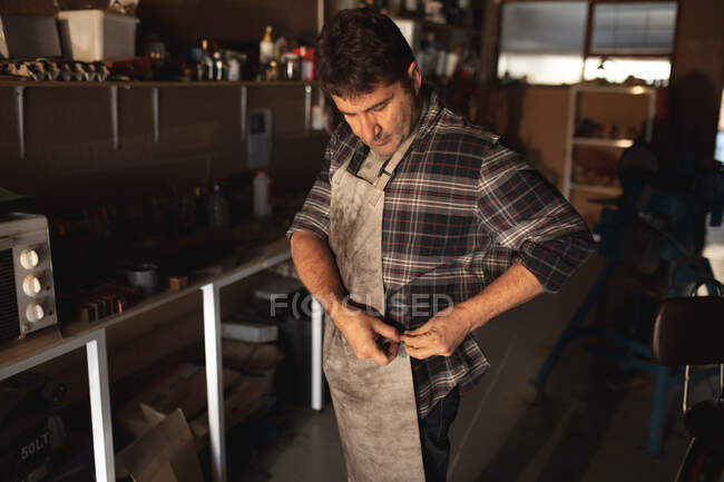 Caucasiano fabricante de facas masculino colocando no avental na oficina. artesão independente de pequenas empresas no trabalho. — Fotografia de Stock