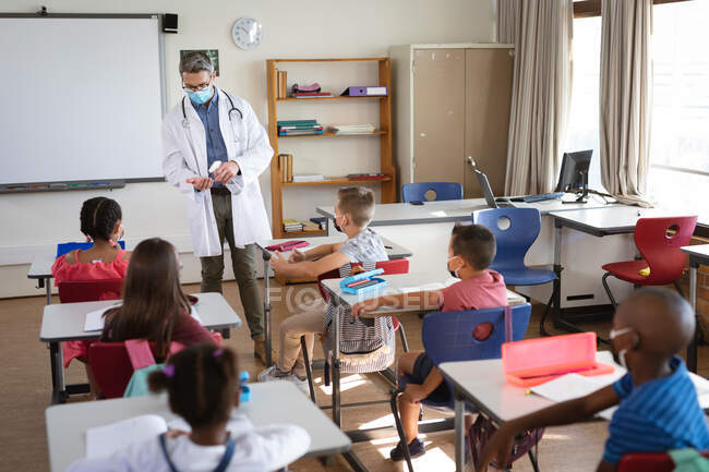 Kaukasischer Arzt mit Mundschutz, der Schülern in der Schule zeigt, wie man Händedesinfektionsmittel anwendet. Gesundheitsschutz und Sicherheit in der Schule während des Covid-19-Pandemiekonzepts — Stockfoto