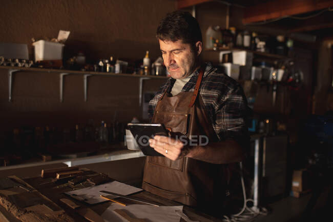 Caucasian male knife maker standing at desk, using tablet in workshop. independent small business craftsman at work. — Stock Photo