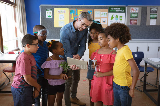 Caucasian male teacher with digital tablet teaching group of diverse students in environment class. school and education concept — Stock Photo