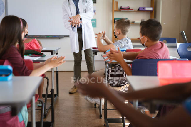 Caucasian male doctor showing how to use hand sanitizer to group of diverse students at school. health protection and safety at school during covid-19 pandemic concept — Stock Photo