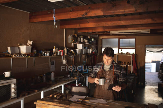 Caucasian male knife maker standing at desk, drinking coffee, using tablet in workshop. independent small business craftsman at work. — Stock Photo