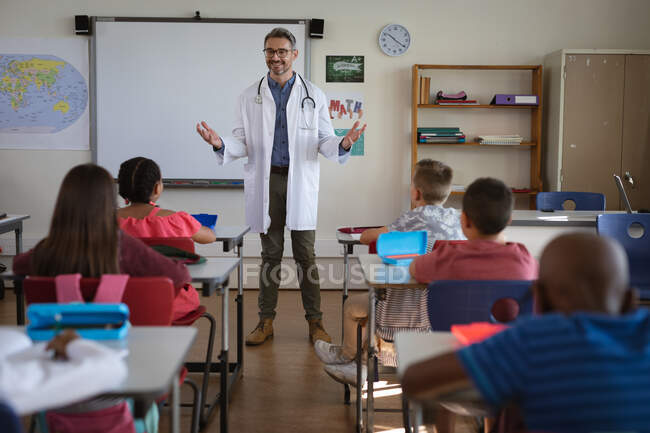 Caucasian male doctor talking to group of diverse students sitting in the class at school. health protection and safety at school during covid-19 pandemic concept — Stock Photo