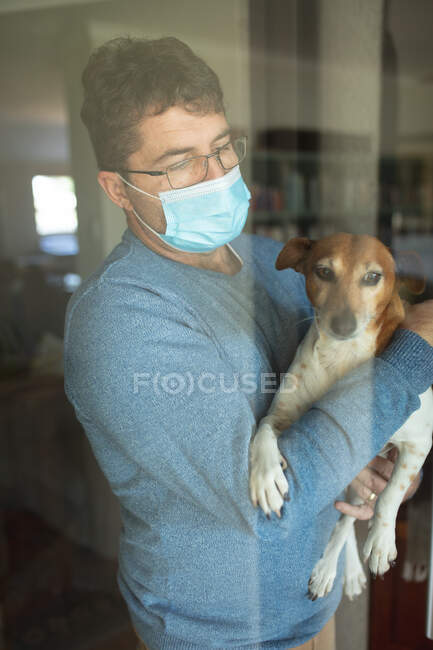 Ritratto di uomo caucasico che indossa occhiali e maschera, tiene il cane, guarda attraverso la finestra. trascorrere del tempo a casa durante la pandemia di coronavirus covid 19. — Foto stock