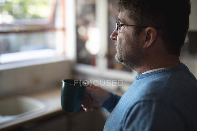 Homem caucasiano focado em pé na cozinha, olhando pela janela, bebendo café. passar tempo livre em casa. — Fotografia de Stock