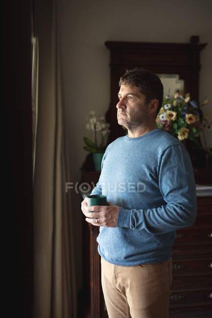 Focused caucasian man standing in living room, looking through window, drinking coffee. spending free time at home. — Stock Photo