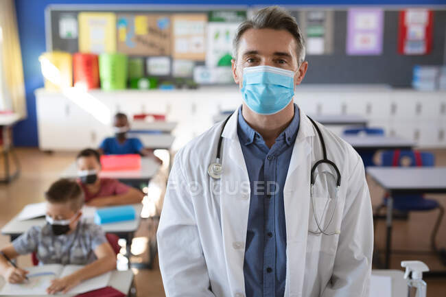 Портрет кавказского врача в маске, стоящей в классе в школе. охрана здоровья и безопасность в школе во время пандемии ковид-19 — стоковое фото