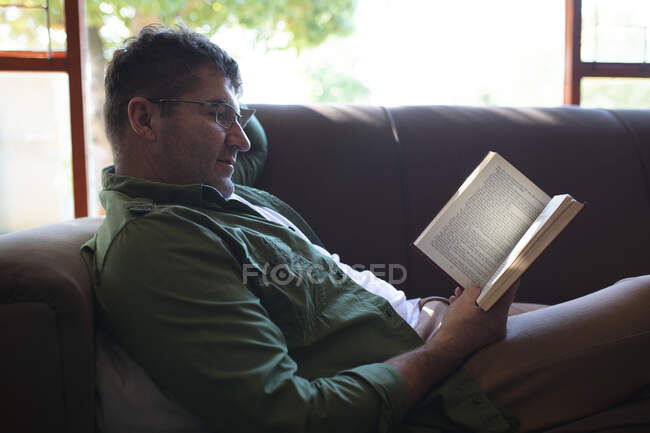 Sonriente hombre caucásico acostado en un sofá, leyendo libro y relajándose. pasar tiempo libre en casa. - foto de stock