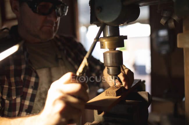 Кавказький чоловічий виробник ножів, одягнений в фартух і окуляри, використовуючи свердло в майстерні. Незалежний дрібний майстер на роботі.. — стокове фото