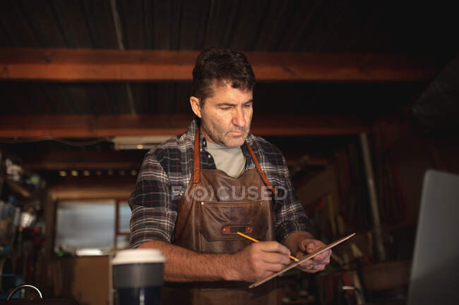 Homem branco fabricante de facas sentado na mesa, fazendo anotações na oficina. artesão independente de pequenas empresas no trabalho. — Fotografia de Stock