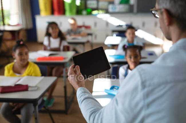 Vista posteriore dell'insegnante maschio caucasico che usa tablet digitale mentre insegna nella classe a scuola. concetto di scuola e istruzione — Foto stock