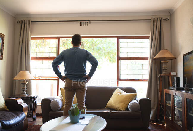 Rückansicht eines kaukasischen Mannes, der im Wohnzimmer steht und aus dem Fenster schaut. Zeit zu Hause verbringen. — Stockfoto