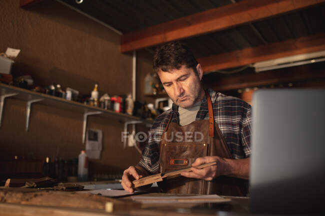 Caucasian male knife maker holding knife, using laptop in workshop. independent small business craftsman at work. — Stock Photo