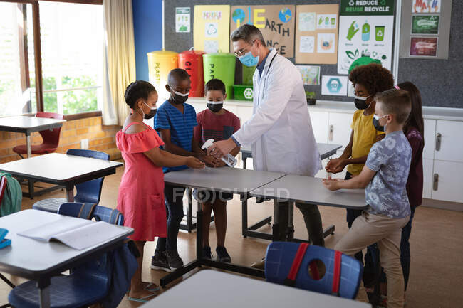 Caucasian male doctor wearing face mask spraying hand sanitizer on hands of students at school. health protection and safety at school during covid-19 pandemic concept — Stock Photo
