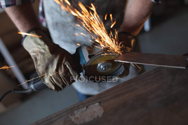 Hände eines männlichen Messermachers mit Schürze, der in der Werkstatt einen Winkelschleifer benutzt. unabhängiger Kleinunternehmer bei der Arbeit. — Stockfoto