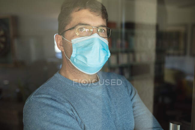Портрет кавказца в очках и маске, смотрящего в окно. Провести время дома во время пандемии коронавируса. — стоковое фото