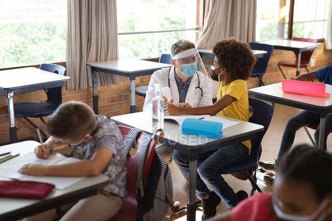 Médecin caucasien portant un écran facial mesurant la température du garçon afro-américain à l'école. protection de la santé et sécurité à l'école pendant la cohabitation 19 concept pandémique — Photo de stock