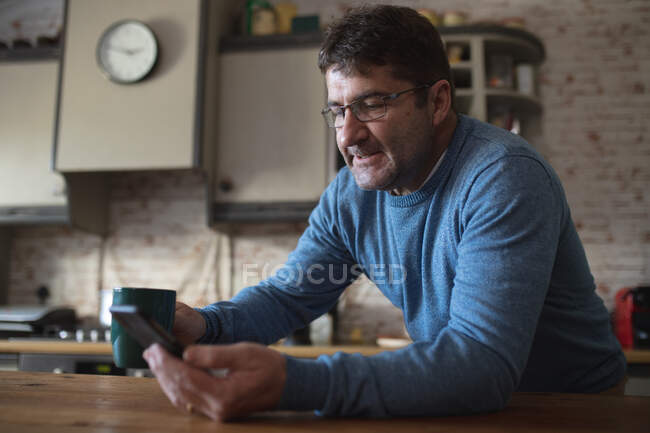 Hombre caucásico en la cocina sentado a la mesa, tomando café y usando un teléfono inteligente. pasar tiempo libre en casa. - foto de stock