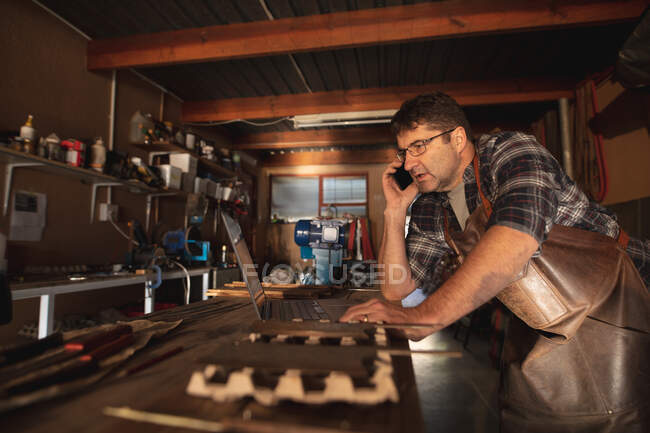 Белый мужчина производитель ножей смартфон и ноутбук в мастерской. независимый ремесленник малого бизнеса за работой. — стоковое фото