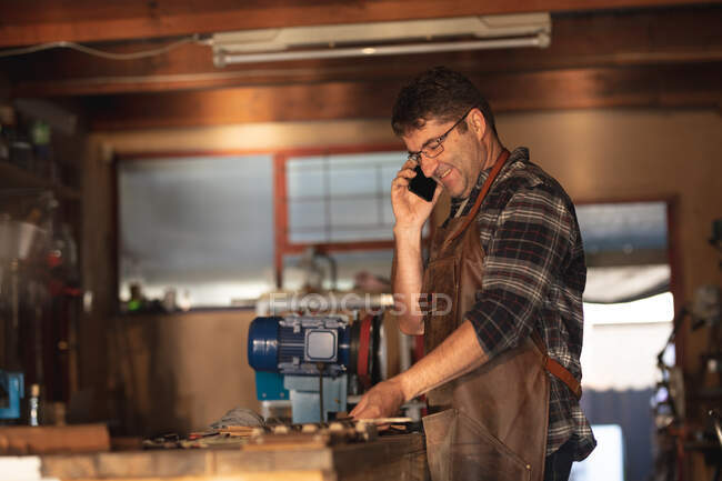Кавказький чоловічий виробник ножів стоїть за столом, розмовляючи на смартфоні в майстерні. Незалежний дрібний майстер на роботі.. — стокове фото