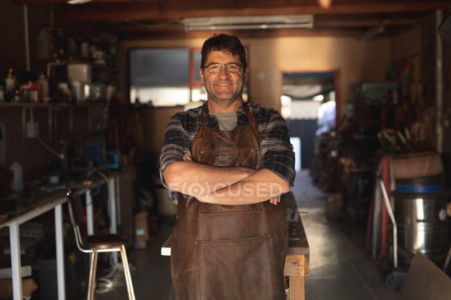 Lächelnder kaukasischer Messermacher mit verschränkten Händen in der Werkstatt, der in die Kamera blickt. unabhängiger Kleinunternehmer bei der Arbeit. — Stockfoto