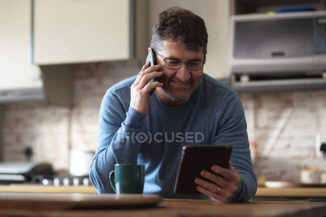 Улыбающийся кавказский мужчина на кухне пьет кофе, пользуется планшетом и смартфоном. проводить свободное время дома. — стоковое фото