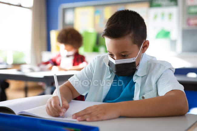 Garçon caucasien portant un masque facial étudiant assis sur son bureau dans la classe à l'école. hygiène et distanciation sociale à l'école pendant la pandémie de covidé 19 — Photo de stock