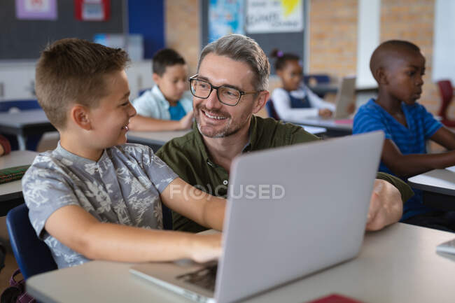Кавказький вчитель чоловічої статі навчає кавказького хлопчика, як користуватися ноутбуком у класі. школа і освіта — стокове фото
