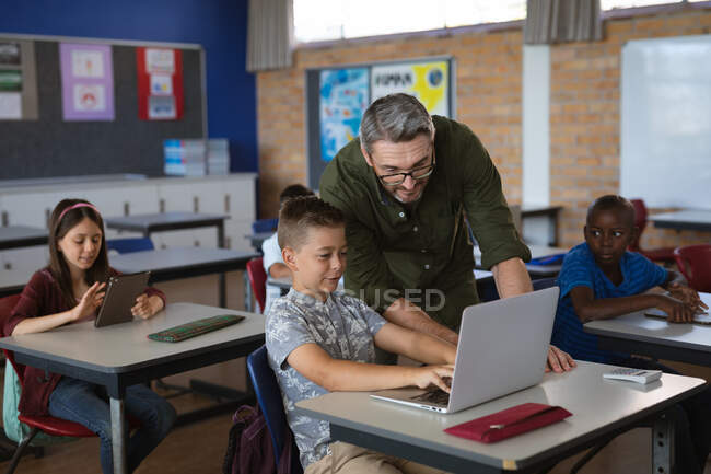 Кавказький вчитель чоловічої статі навчає кавказького хлопчика, як користуватися ноутбуком у класі. школа і освіта — стокове фото