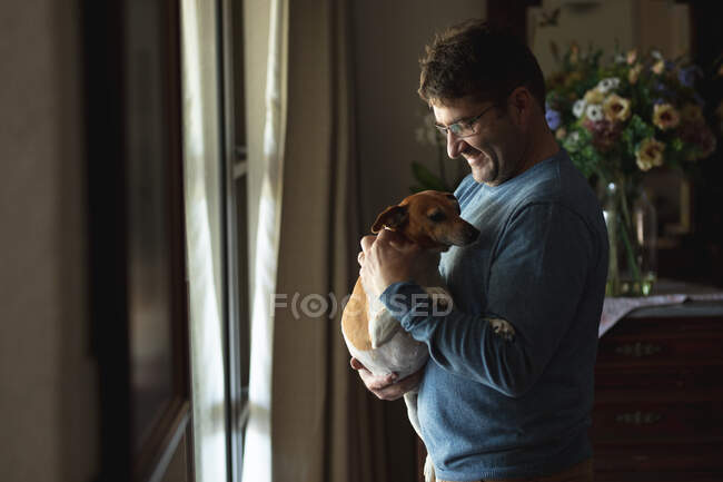 Lächelnder Kaukasier, der am Fenster im Wohnzimmer steht und Hund streichelt. Freizeit zu Hause verbringen. — Stockfoto