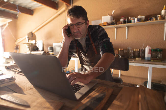 Coltellinaio maschio caucasico smartphone e laptop in officina. artigiano indipendente delle piccole imprese al lavoro. — Foto stock