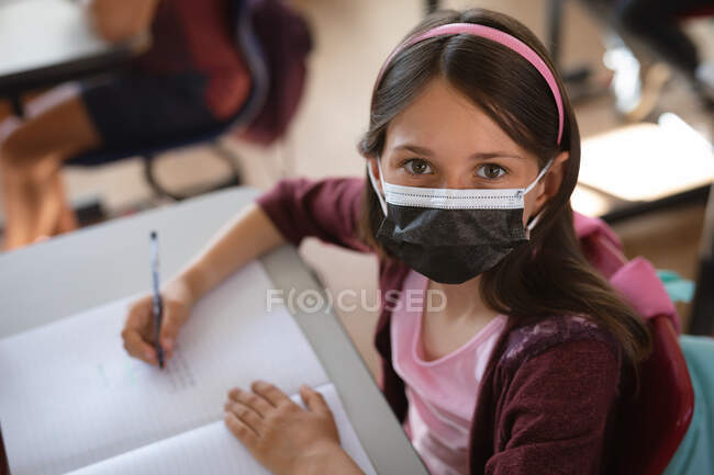 Retrato de una chica caucásica con máscara facial sentada en su escritorio en la clase de la escuela. higiene y distanciamiento social en la escuela durante la pandemia de covid 19 - foto de stock