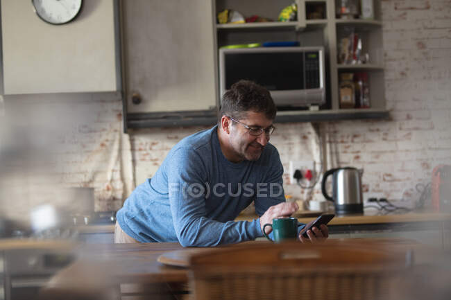 Улыбающийся кавказский мужчина на кухне, стоящий за столом, пьющий кофе и пользующийся смартфоном. проводить свободное время дома. — стоковое фото